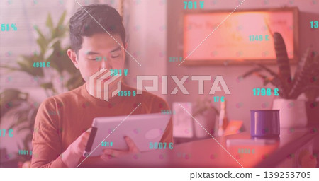 Chinese man wearing brown shirt tapping tablet at desk in home office, floating currency symbols 139253705