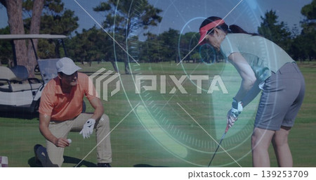 Kneeling golfer in orange polo placing ball on fairway, partner in green-polo swinging club by cart 139253709
