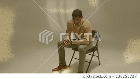 Leaning forward man wearing beige sweater sitting on black folding chair against grey backdrop Leaning forward man wearing beige sweater sitting on black folding chair against grey backdrop 139253727