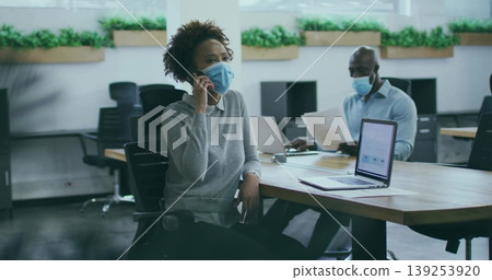 Woman holding smartphone wearing mask in open-plan office, with coworker reading papers at laptops Woman holding smartphone wearing mask in open-plan office, with coworker reading papers at laptops 139253920