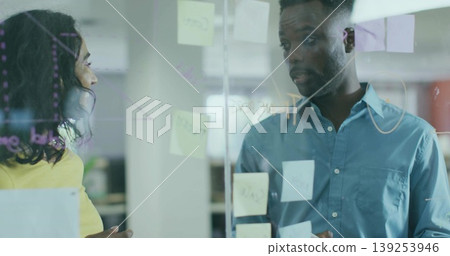 Collaborating pair in yellow blouse blue shirt at glass board with sticky notes in open office 139253946
