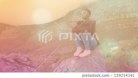 Sitting African American woman in sweater on rock boulder on hill gazing toward sunset, copy space 139254191