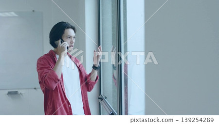 Checking smartphone man wearing red shirt by large glass window in office, with whiteboard Checking smartphone man wearing red shirt by large glass window in office, with whiteboard 139254289