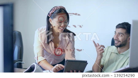 Woman in pink shirt leaning over desk presenting tablet screen to colleague in office, with phone 139254290