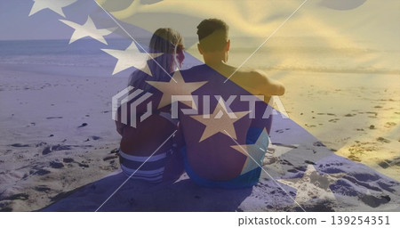 Sitting couple wearing swimwear gazing toward horizon at sandy beach waters edge, with flag overlay Sitting couple wearing swimwear gazing toward horizon at sandy beach waters edge, with flag overlay 139254351