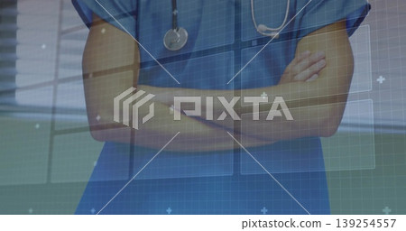 Standing male doctor crossing arms in corridor wearing scrubs, stethoscope with digital interface Standing male doctor crossing arms in corridor wearing scrubs, stethoscope with digital interface 139254557