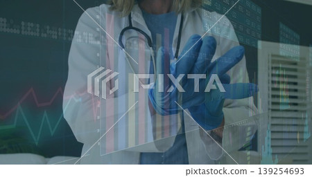 Provider in white coat blue scrubs and stethoscope pulling on gloves in clinic with digital charts 139254693