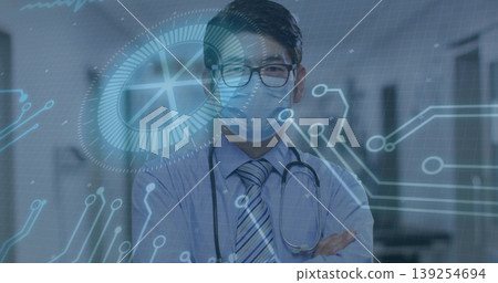 Standing doctor crossing arms in hospital corridor, with mask, specs, stethoscope, hologram overlay 139254694