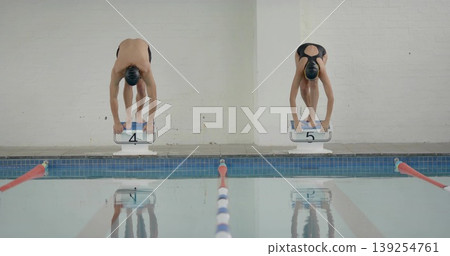 Two swimmers wearing swim caps, suits bending forward on starting blocks in pool, with lane ropes Two swimmers wearing swim caps, suits bending forward on starting blocks in pool, with lane ropes 139254761