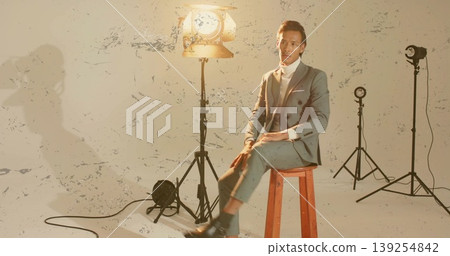 Asian man wearing grey suit sitting on stool in studio, with lights, cords on paint-splattered wall Asian man wearing grey suit sitting on stool in studio, with lights, cords on paint-splattered wall 139254842