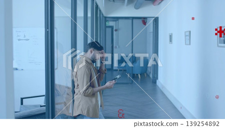Walking Indian man wearing shirt and jeans holding tablet in glass corridor, with whiteboard Walking Indian man wearing shirt and jeans holding tablet in glass corridor, with whiteboard 139254892