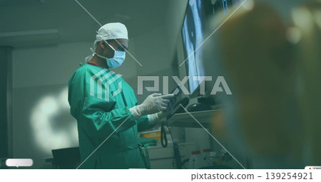 Operating green-scrubbed masked gloved surgeon touching interface in OR with monitors and cart Operating green-scrubbed masked gloved surgeon touching interface in OR with monitors and cart 139254921