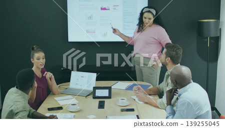 Hispanic woman pointing at display in conference room, with laptops mugs tablet phone, copy space Hispanic woman pointing at display in conference room, with laptops mugs tablet phone, copy space 139255045