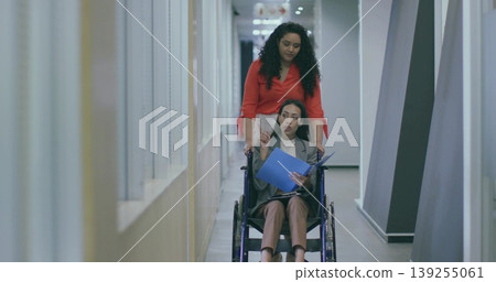 Woman in red blouse pushing colleague in grey blazer wheelchair down corridor, holding blue folder Woman in red blouse pushing colleague in grey blazer wheelchair down corridor, holding blue folder 139255061