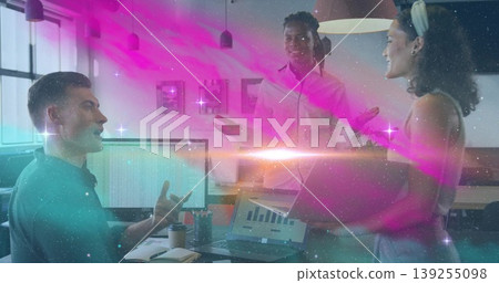 Gesturing seated man pointing at monitors in office, with laptop, notebook and cosmic light overlay 139255098