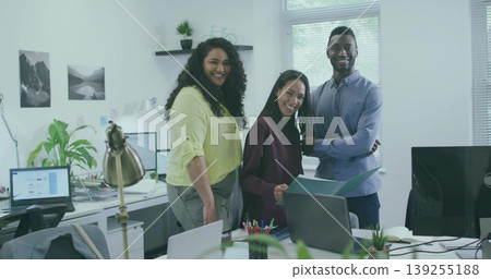 Smiling three coworkers wearing work attire standing at workstation in office, with laptop monitors 139255188