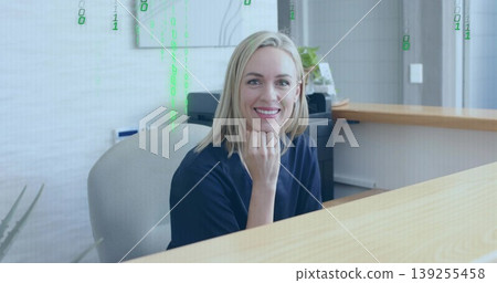 Smiling receptionist wearing navy blouse at reception desk, with printer and green code overlay 139255458