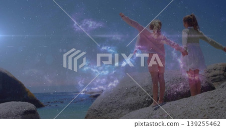 Standing sisters holding hands amid purple nebula glowing sparks on coastal boulders, copy space Standing sisters holding hands amid purple nebula glowing sparks on coastal boulders, copy space 139255462