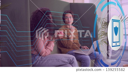 Two colleagues analyzing circuit and shield overlays on green lounge sofa with red mug and laptop 139255582