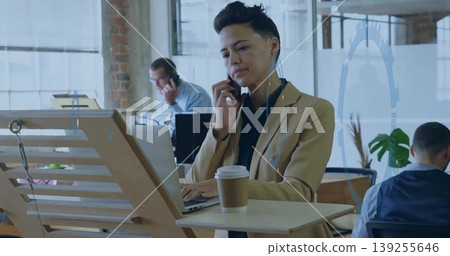 Holding smartphone, Asian woman in blazer typing on laptop at desk in office, with coffee cup Holding smartphone, Asian woman in blazer typing on laptop at desk in office, with coffee cup 139255646
