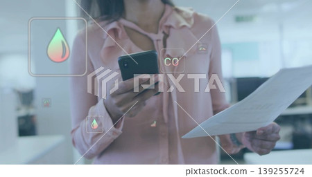 Female office worker analyzing overlay in open-plan office, with smartphone, document and CO2 icons 139255724