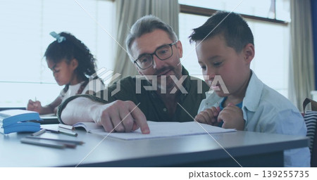 Pointing male tutor guiding boy student at study table near window with pencils and sharpener Pointing male tutor guiding boy student at study table near window with pencils and sharpener 139255735