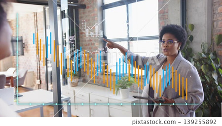 Woman wearing blazer pointing at charts on glass board in open-plan office, with tablet, copy space Woman wearing blazer pointing at charts on glass board in open-plan office, with tablet, copy space 139255892