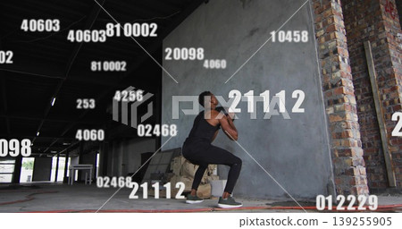 Squatting man wearing tank top holding kettlebell inside warehouse, with debris and brick pillars Squatting man wearing tank top holding kettlebell inside warehouse, with debris and brick pillars 139255905