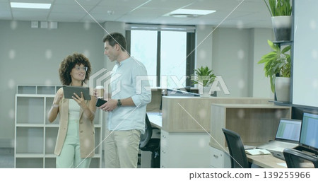 Gesturing woman in blazer and man discussing tablet at open-plan office, with coffee cup smartphone 139255966