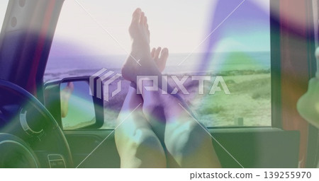 Resting bare legs crossing ankles on open car window sill, with steering wheel and sandy shoreline Resting bare legs crossing ankles on open car window sill, with steering wheel and sandy shoreline 139255970