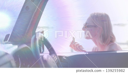 Leaning woman applying lip balm through passenger window at waterfront with lens flare copy space Leaning woman applying lip balm through passenger window at waterfront with lens flare copy space 139255982