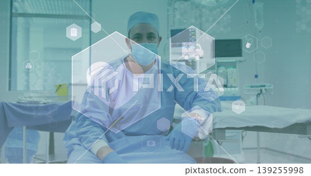 Sitting male surgeon wearing blue scrubs with mask and gloves in surgical suite with monitors Sitting male surgeon wearing blue scrubs with mask and gloves in surgical suite with monitors 139255998