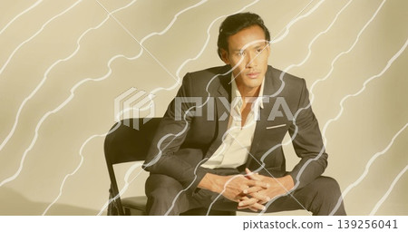 Leaning Asian man wearing dark gray suit clasping hands on chair in studio, with abstract overlay 139256041