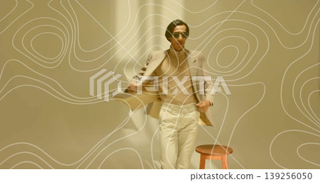Standing Asian male model wearing beige blazer and trousers in minimalist studio, with wooden stool 139256050