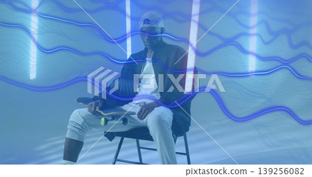 Holding skateboard skateboarder sitting on stool in studio, with neon tubes and blue wave trails Holding skateboard skateboarder sitting on stool in studio, with neon tubes and blue wave trails 139256082