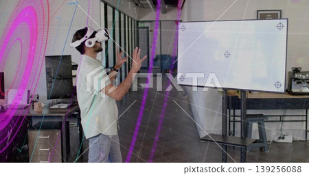 Interacting man wearing VR headset gesturing toward wall-mounted screen in open-plan office 139256088