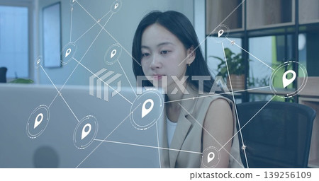 Typing woman wearing blazer using laptop at office desk with potted plant, viewing location pins 139256109