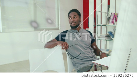 Leaning man holding glasses, looking off to side at glass desk in office, with monitor, books Leaning man holding glasses, looking off to side at glass desk in office, with monitor, books 139256275