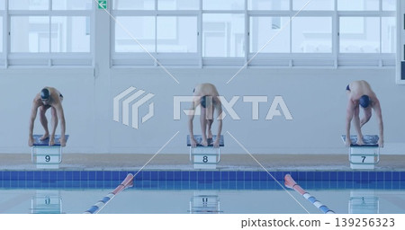 Preparing three male swimmers gripping starting blocks on competition pool deck, with windows Preparing three male swimmers gripping starting blocks on competition pool deck, with windows 139256323