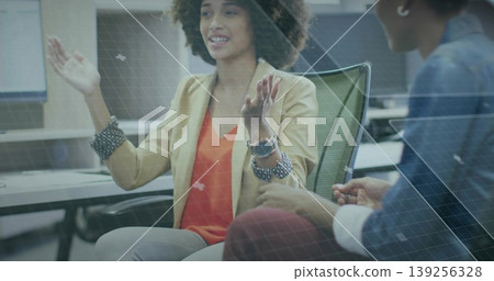 Gesturing woman wearing beige blazer discussing at office, with green mesh chairs and monitors 139256328