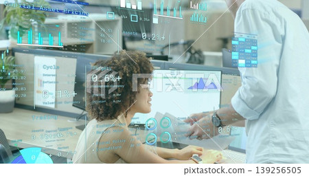 Seated woman and standing coworker reviewing financial charts on monitors in office, with documents Seated woman and standing coworker reviewing financial charts on monitors in office, with documents 139256505