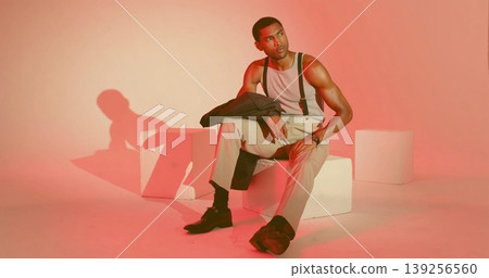 Man wearing tank top with suspenders gazing while seated on cube in peach studio, holding jacket Man wearing tank top with suspenders gazing while seated on cube in peach studio, holding jacket 139256560