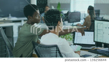 Two adult women collaborating in office, pointing at blueprint on monitor with laptops coffee cup 139256644