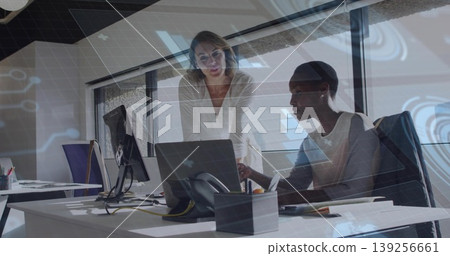 Collaborating two female colleagues reviewing laptop screen at corporate office, with notepads Collaborating two female colleagues reviewing laptop screen at corporate office, with notepads 139256661