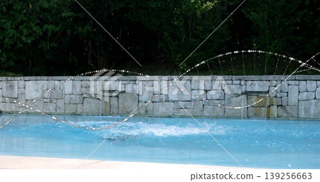 Arcing water jets splashing blue water at backyard pool, with stone wall fountains, concrete deck Arcing water jets splashing blue water at backyard pool, with stone wall fountains, concrete deck 139256663