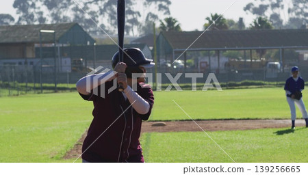 Swinging batter wearing maroon jersey black helmet gripping bat on diamond, with protective fence Swinging batter wearing maroon jersey black helmet gripping bat on diamond, with protective fence 139256665