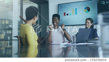 Celebrating woman in blouse raising fists at table in glass meeting room, with sales tracker screen Celebrating woman in blouse raising fists at table in glass meeting room, with sales tracker screen 139256834