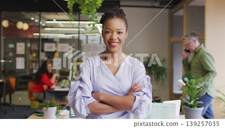 Smiling Black woman wearing striped blouse crossing arms in open office, with laptop and smartphone Smiling Black woman wearing striped blouse crossing arms in open office, with laptop and smartphone 139257035