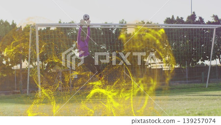 Jumping lone goalkeeper catching soccer ball above goalpost on pitch, with gloves and yellow light 139257074