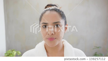 Looking Indian woman wearing white bathrobe in bright room, with silver nose ring and green plants 139257112